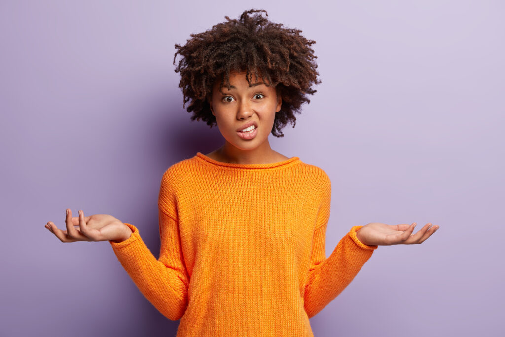 Attractive woman with curly hair and an orange sweater looking confused against a light purple background.